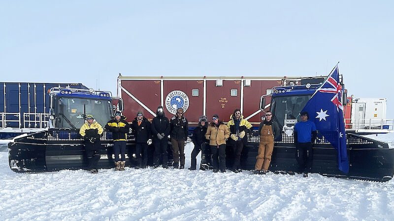 A group of people stand on the snow in front of the tractor train