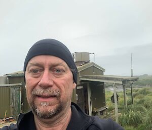 A man in a black beanie takes a selfie in front of an old green hut on a grey day