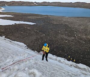 A man in a yellow jacket and blue helmet abseils down a snow-covered cliff. A group of people in yellow outfits can be seen at the bottom of the cliff. In the background there is a blue lake and rocky hills.