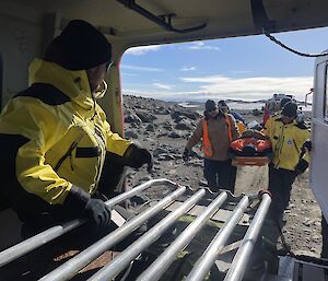 Four men loading a stretcher with a person on it into the back of a rescue Hägglund. There is a person in the back of the vehicle ready to assist with the stretcher.