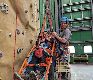 A smiling man is suspended from a climbing wall with abseiling ropes and is supporting a smiling woman in an almost horizontal orange stretcher who is also suspended off the climbing wall with abseiling ropes.