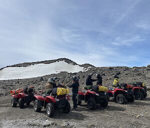 Five red quad bikes with riders standing next to them. The background is sand and rock with a snow covered hill visible in the distance.