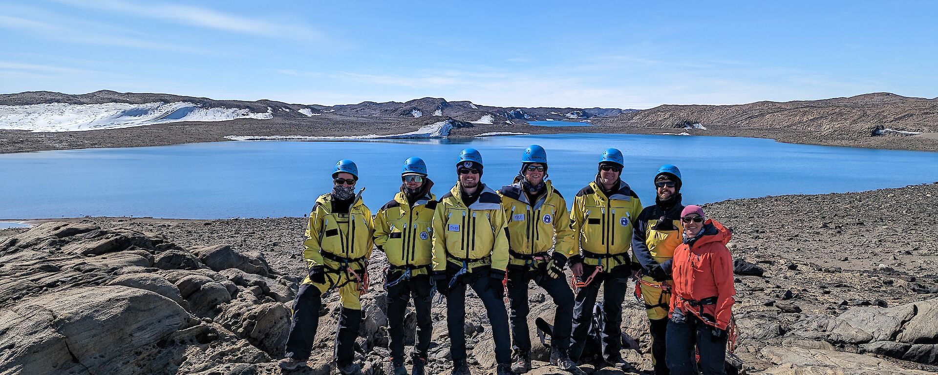 A group of six smiling men wearing yellow jackets and blue helmets and one smiling woman wearing an orange jacket all stand together on a rocky outcrop with a blue lake in the distant background.