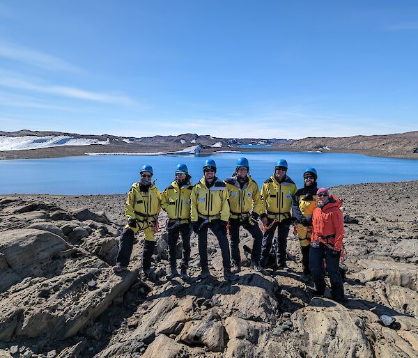 A group of six smiling men wearing yellow jackets and blue helmets and one smiling woman wearing an orange jacket all stand together on a rocky outcrop with a blue lake in the distant background.