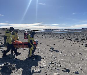 Four people in yellow outfits carry an orange stretcher with a person in it over rocky terrain.