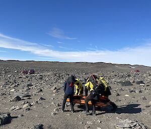 Four stretcher bearers in rocky terrain are about two to three hundred metres away from two rescue vehicles (Hägglunds)