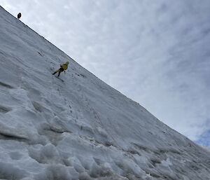 A man in a yellow outfit has abseiled half-way down a snow-covered cliff-face. The picture is taken looking up towards the cloudy sky and another man can be seen at the top of the icy cliff looking downward.