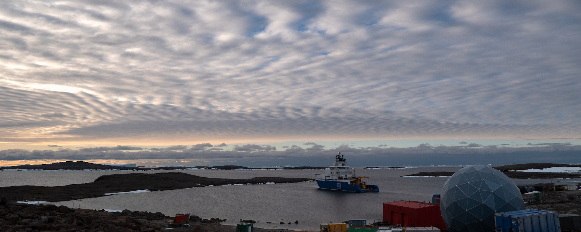 A blue and white ship sits calmly in a natural harbour under wispy white clouds