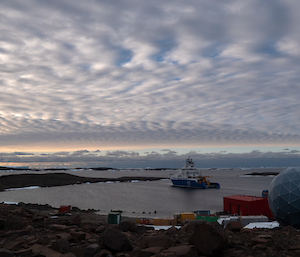 A blue and white ship sits calmly in a natural harbour under wispy white clouds