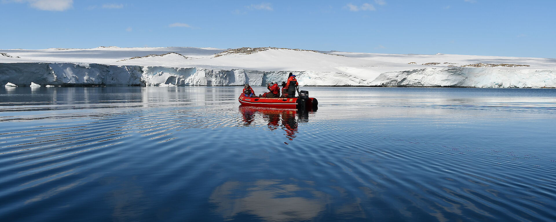A red inflatable boat with five people in the middle of a blue bay surrounded by white ice cliffs.