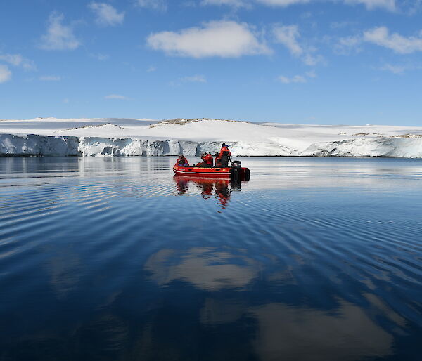 A red inflatable boat with five people in the middle of a blue bay surrounded by white ice cliffs.