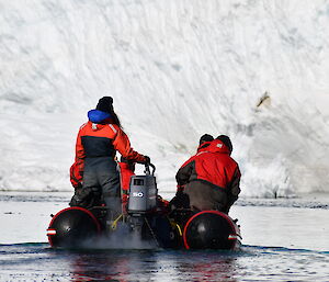 View of an inflatable boat from behind with five people, being driven towards a towering ice cliff.