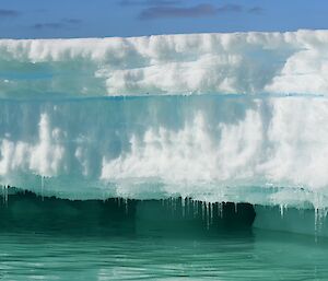 A multi-shade iceberg with an overhang of icicles almost touching turquoise water with a blue sky.