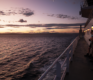 A small group of people on the deck of a ship looking at the sunset over land