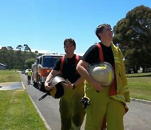 A man and a woman walking on a road in yellow fire-fighting overalls holding helmets. Red fire truck in the background.