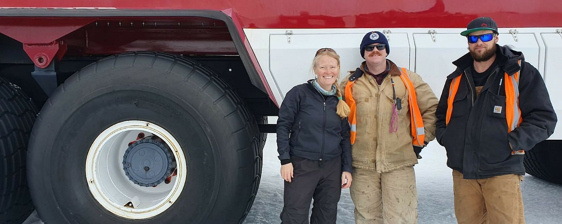 Three people standing in front of large red and white bus, with the bus wheel on the left being a similar height to the people in the photo.
