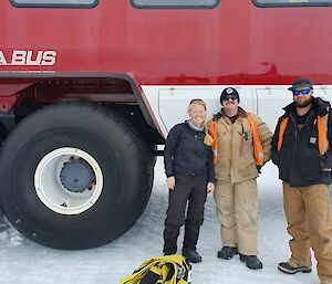 Three people standing in front of large red and white bus, with the bus wheel on the left being a similar height to the people in the photo.