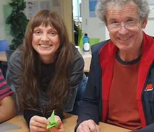 Two people smiling at the camera from behind a table. Person on the left holding a green origami crane in their hand.