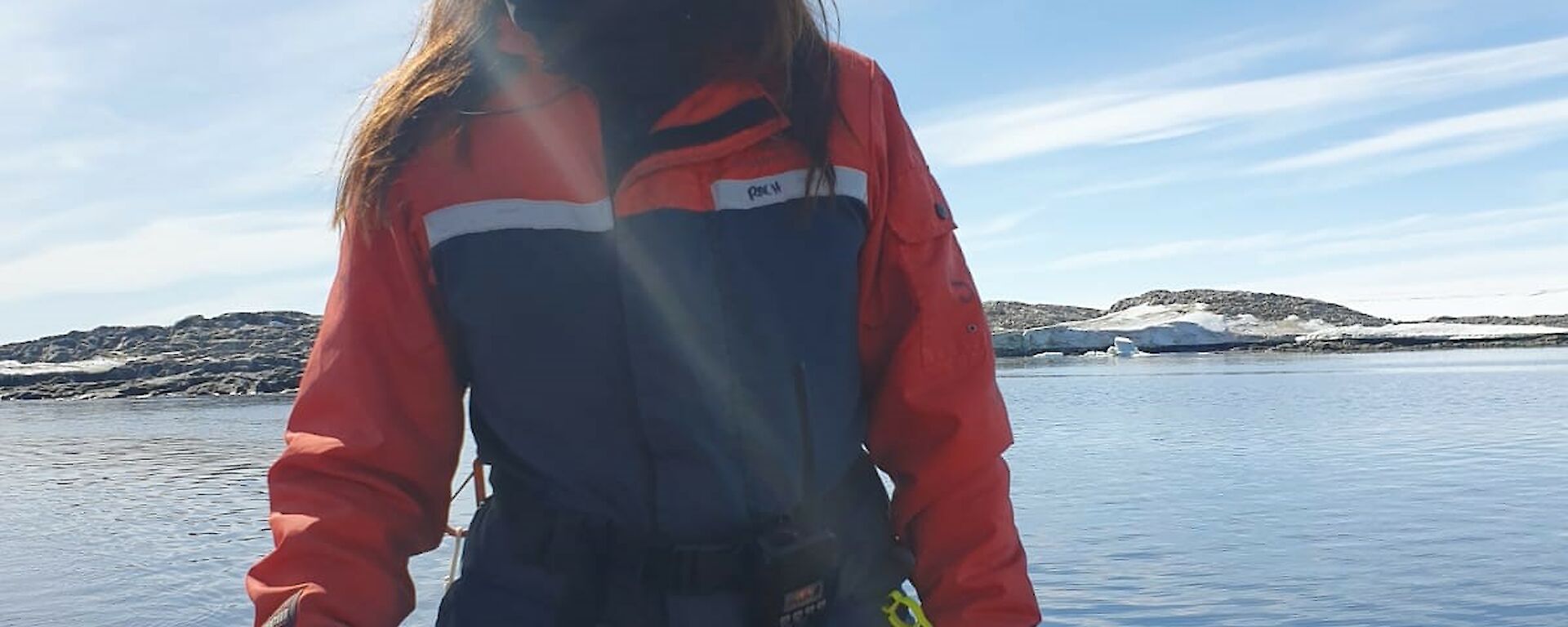 Woman smiling and kneeling on inflatable boat holding tiller. Blue water and snow-covered rocks in the background, with sun shining.