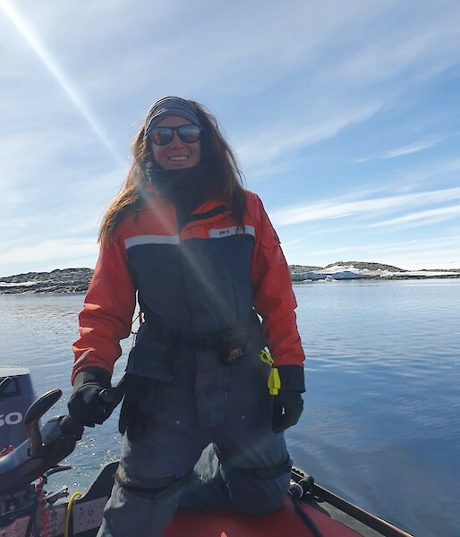 Woman smiling and kneeling on inflatable boat holding tiller. Blue water and snow-covered rocks in the background, with sun shining.