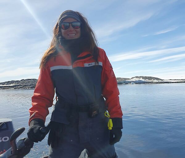 Woman smiling and kneeling on inflatable boat holding tiller. Blue water and snow-covered rocks in the background, with sun shining.