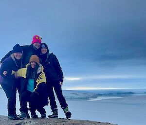Four people pose on a rock in front of a bay with ice cliffs in the background and cloudy skies.