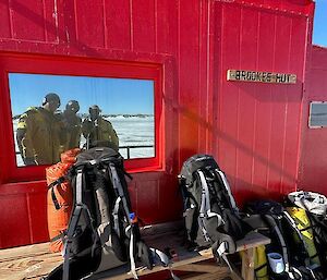 A red field hut with a Brookes Hut sign on the front. There are three field packs sitting out the front of the hut and the reflection of three expeditioners can be seen in the front window of the hut.