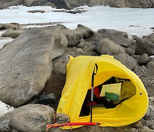 A yellow bivvy bag propped open with a walking pole. The bivvy is sitting on rocky ground between boulders with snow and rocky hills in the background.