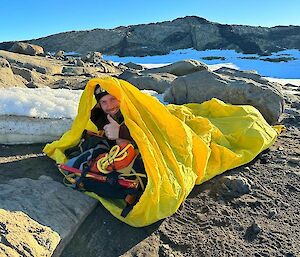 A smiling man with the thumbs up sign lies inside a yellow bivvy bag on rocky ground with snow in the background.