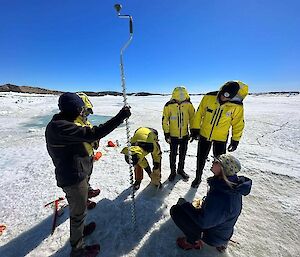 A group of six people standing on sea-ice learn how to use a large 2.5m high sea-ice hand-drill.