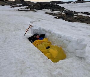 A smiling man lying inside a yellow bivvy bag inside a snow burrow. The windy side of the snow burrow is sheltered by a wall, about 80cm high, built of snow-bricks.