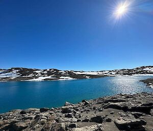A blue lake under the bright sunlight surrounded by rocky shores with snow covered hills in the distance.