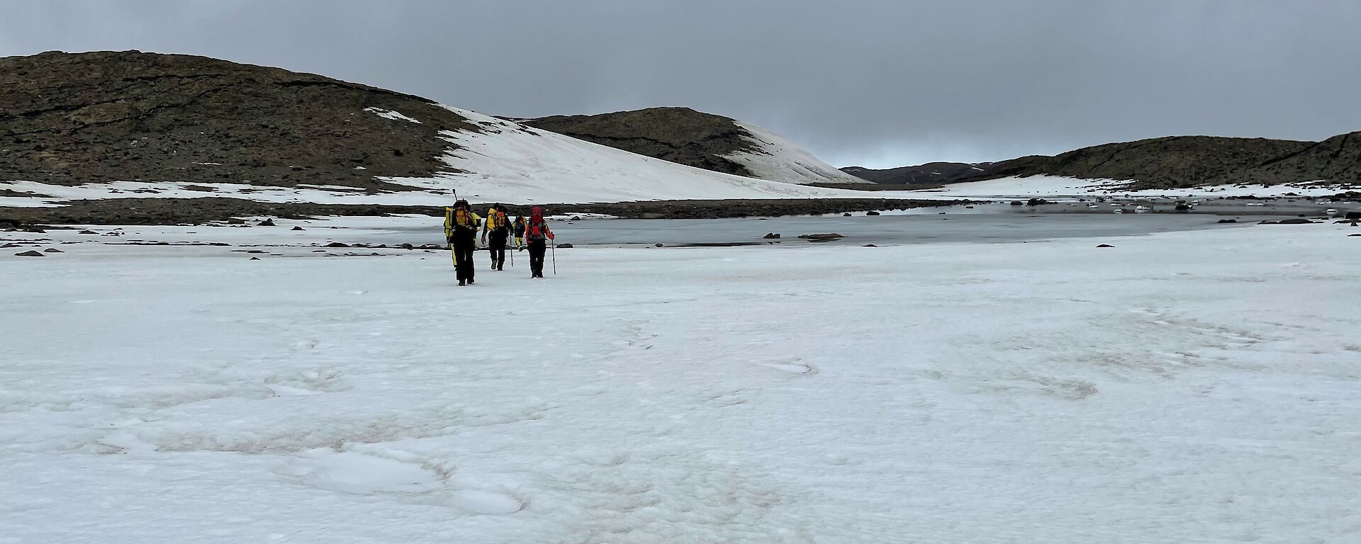 A group of three people in the distance wearing backpacks hiking across the snow towards an icy lake.