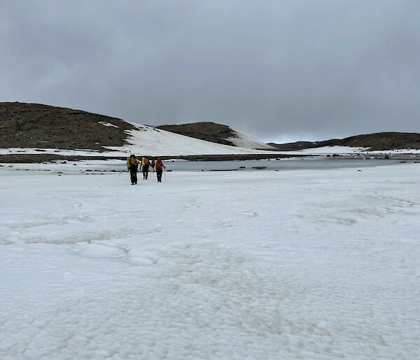 A group of three people in the distance wearing backpacks hiking across the snow towards an icy lake.