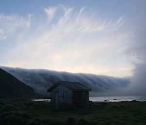 A line of low cloud rolls over an island with a small wooden hut