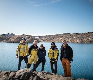 Five smiling people in warm outdoor clothing standing in front of a lake with rocky hills in the background..