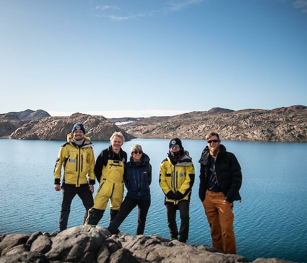 Five smiling people in warm outdoor clothing standing in front of a lake with rocky hills in the background..
