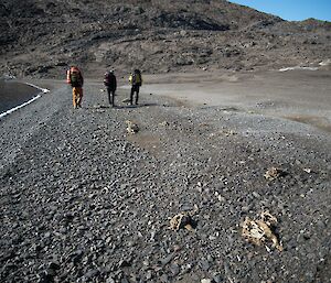 A group of three people walking along the shores of a lake. A rocky hill can be seen in the distance.