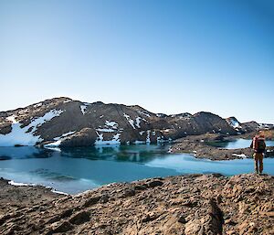 A man with a backpack stands on a rocky hill and looks over a semi-frozen lake. There are snow covered hills in the distance.