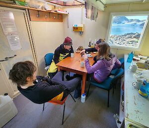 A group of four people sitting around a table playing a game of cards. There is a view of a lake and rocky hills through the window that is in front of the table.