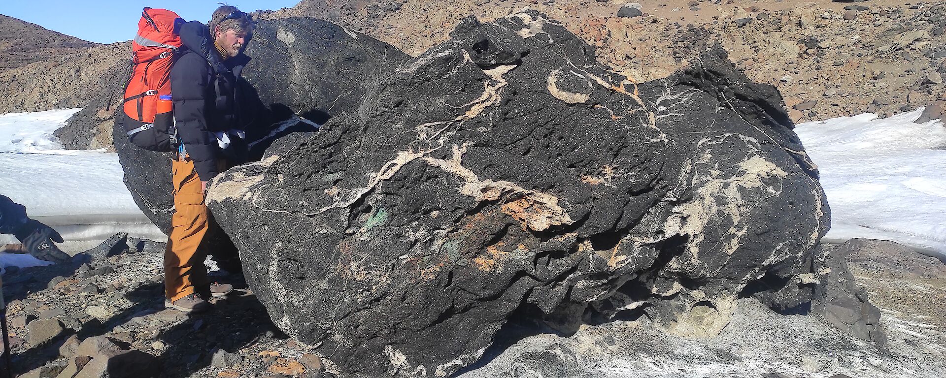 A man wearing a field pack standing next to a black and white swirled rock. The rock is about five times his size.