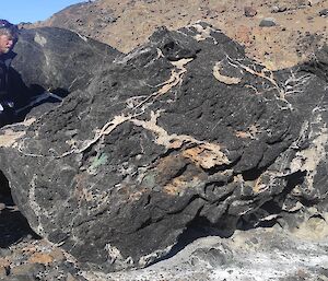 A man wearing a field pack standing next to a black and white swirled rock. The rock is about five times his size.