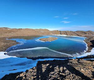 A semi-frozen lake showing lots of different colours. The lake is surrounded by rocky hills and in the distance there are long lines of black rock cutting through the hills.