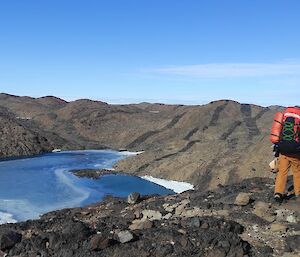 A group of three people wearing backpacks stands at the top of a rocky hill overlooking a distant lake also surrounded by rocky hills.