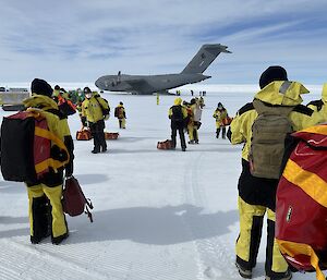 A crowd of people in Antarctic survival gear look on towards a C-17A in the background.