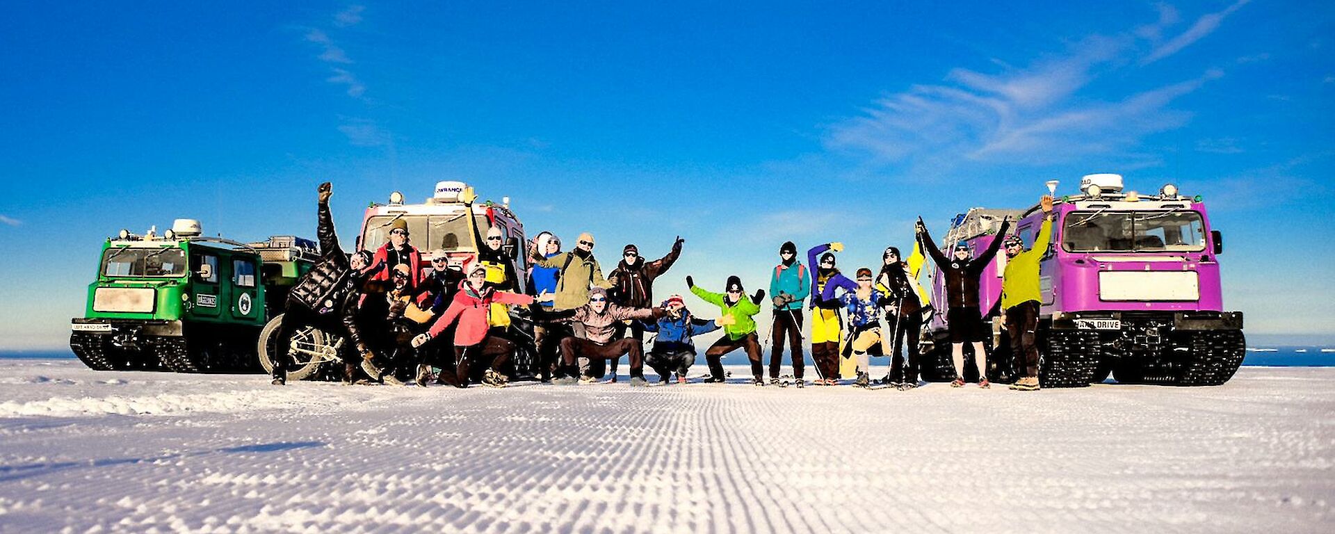 A dozen people in Antarctic running gear pose with arms raised in front of colourful Hagglunds