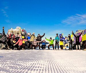 A dozen people in Antarctic running gear pose with arms raised in front of colourful Hagglunds