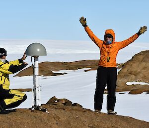 On an Antarctic hill, a scientist works on an instrument dome while an FTO in a bright orange jacket raises her arms in celebration