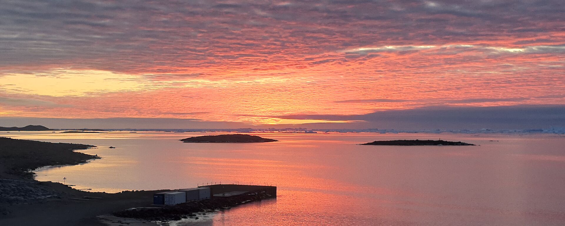 The still waters of a calm bay reflect the pink-red skies overhead.