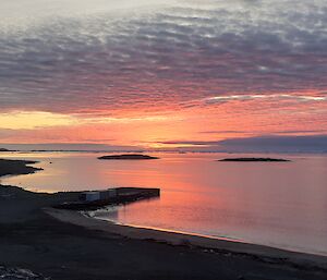 The still waters of a calm bay reflect the pink-red skies overhead.